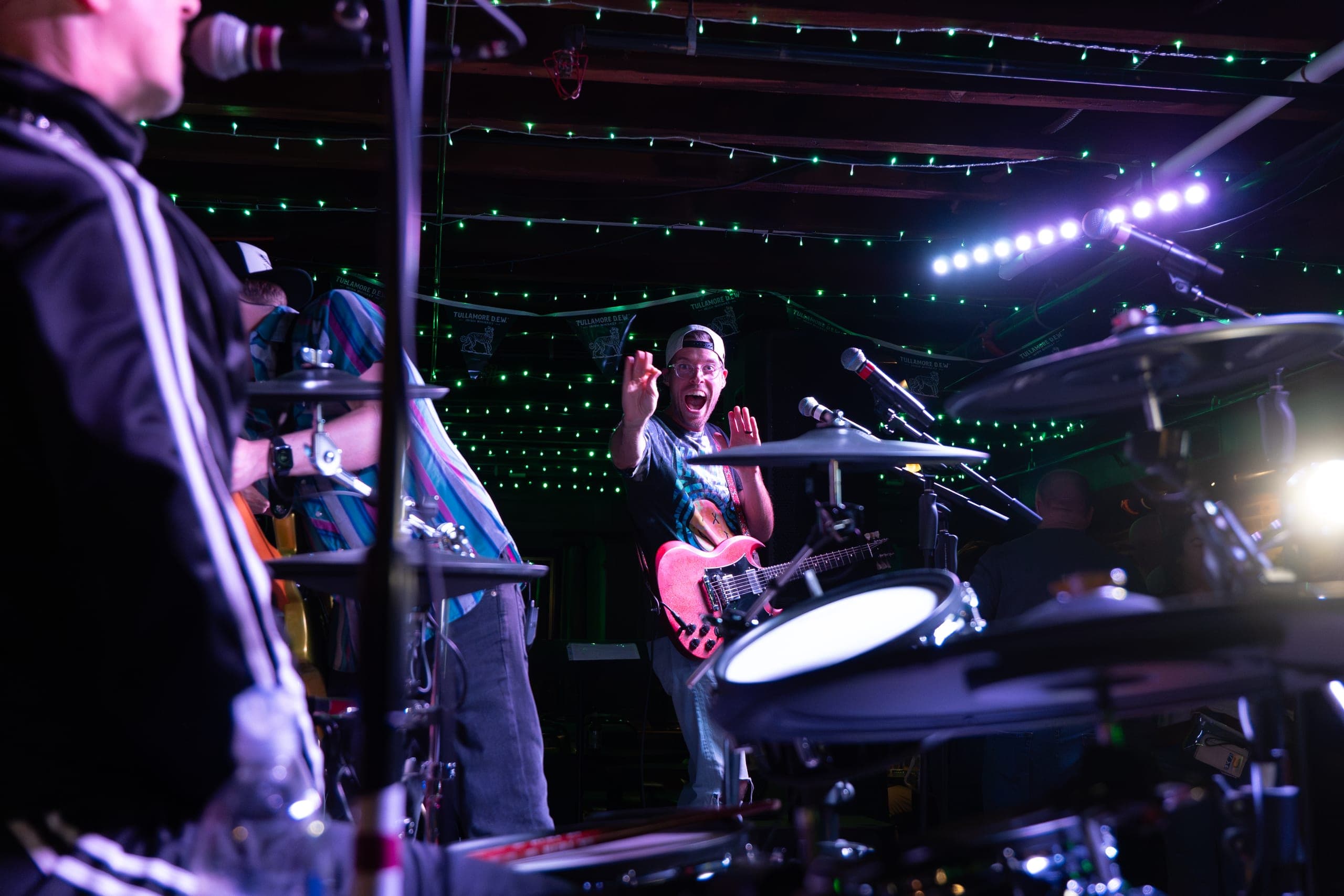 Guitarist with a pink SG flashing an excited grin, seen from behind the drum kit